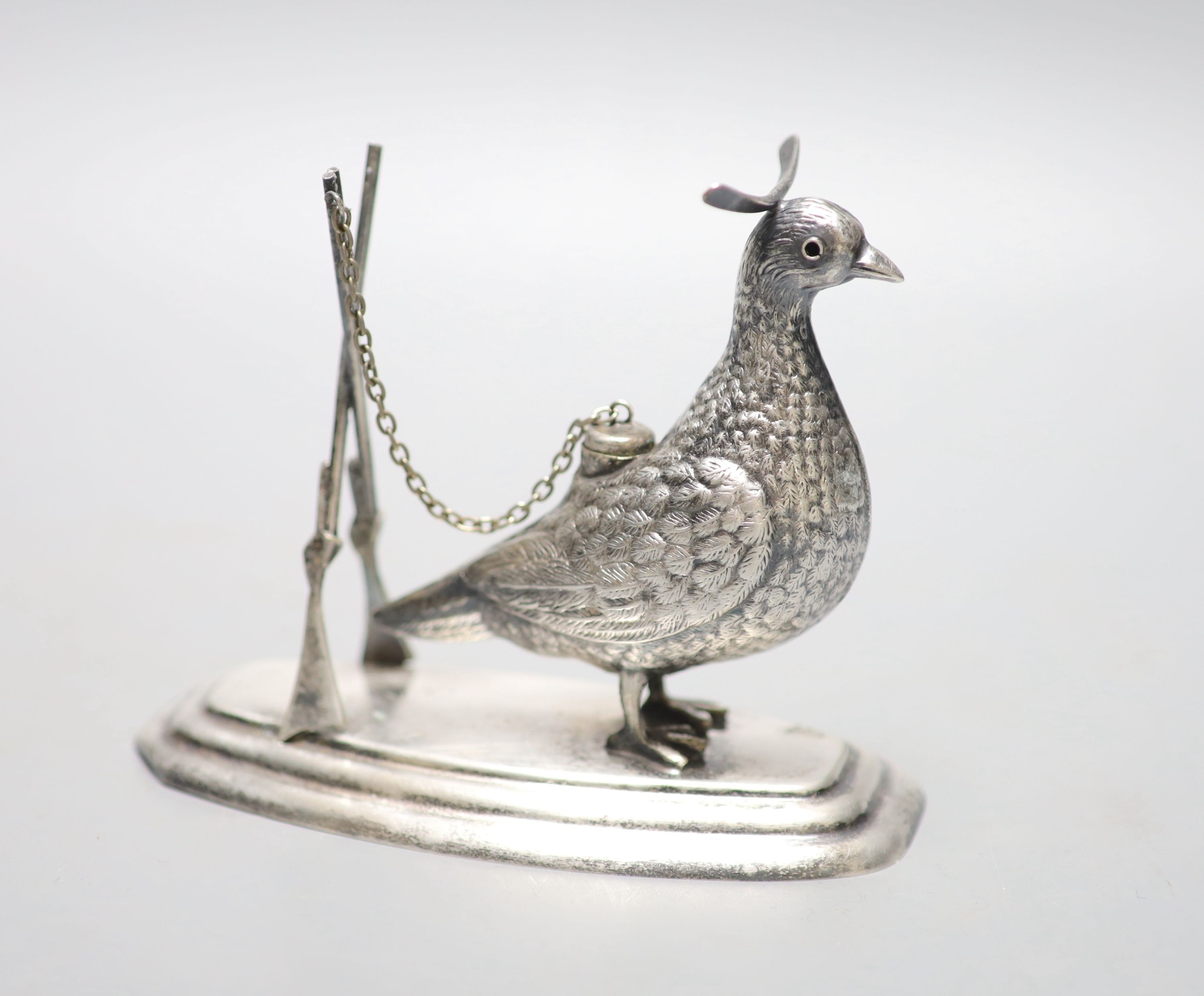 A continental novelty white metal brand balloon warmer, modelled and a game bird with crossed rifles, on a shaped oval base, 13.1cm, indistinct stamp.